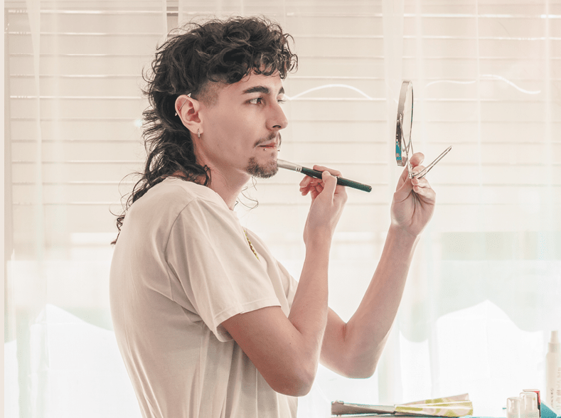 Young non-binary make-up artist applying makeup in front of a mirror with light bulbs in a bright room, focusing on diversity and inclusion in the beauty industry